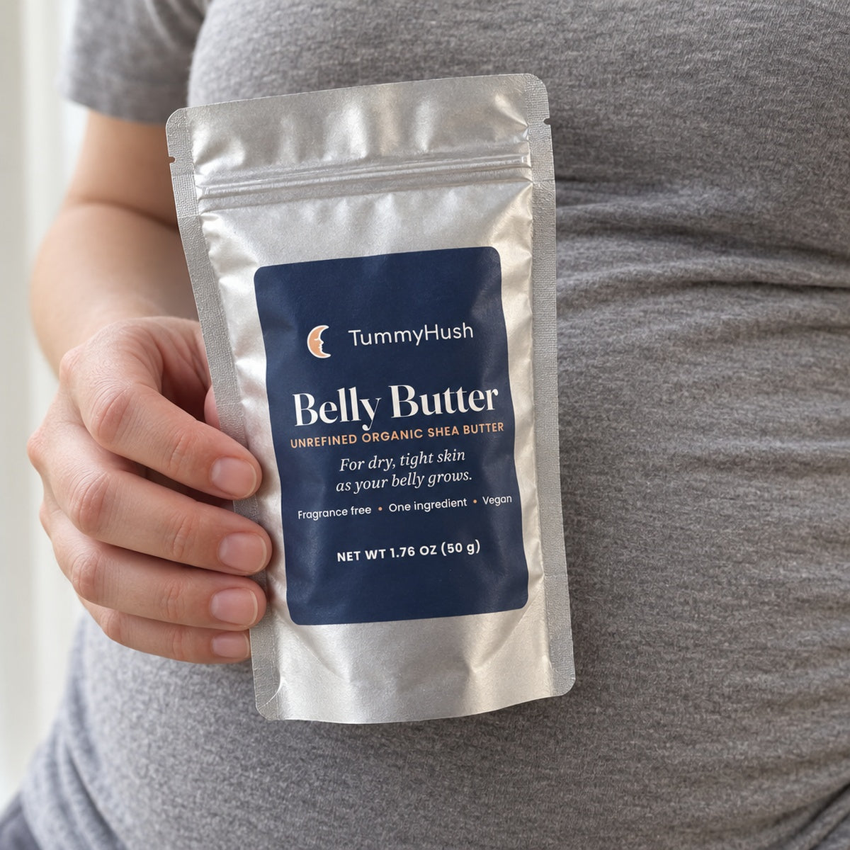 TummyHush Belly Butter pouch held in one hand against a grey shirt