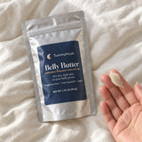 TummyHush Belly Butter pouch on bedding beside a fingertip swipe of shea butter